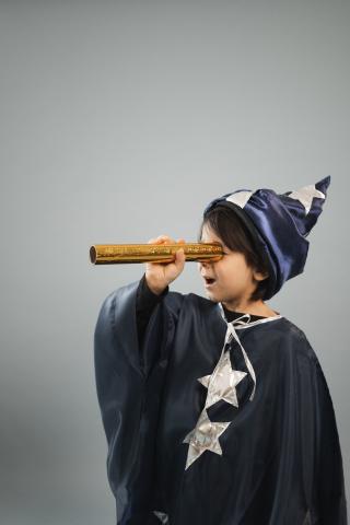 A young child wearing magician robes looks excitedly into a kaleidoscope tube.